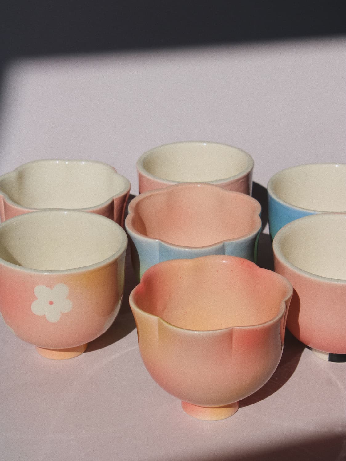 A set of pastel ceramic tea cups arranged on a light surface, featuring soft pink, peach, light blue, and cream colors with a white flower motif on one cup.