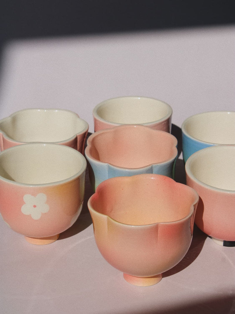 A set of pastel ceramic tea cups arranged on a light surface, featuring soft pink, peach, light blue, and cream colors with a white flower motif on one cup.