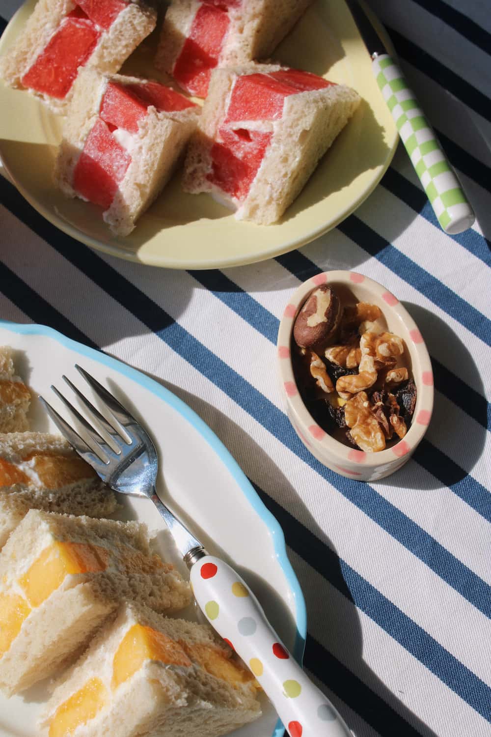 A styled food scene with sliced sandwich bread pieces, a small bowl of nuts, and patterned tableware on a blue-and-white striped cloth.