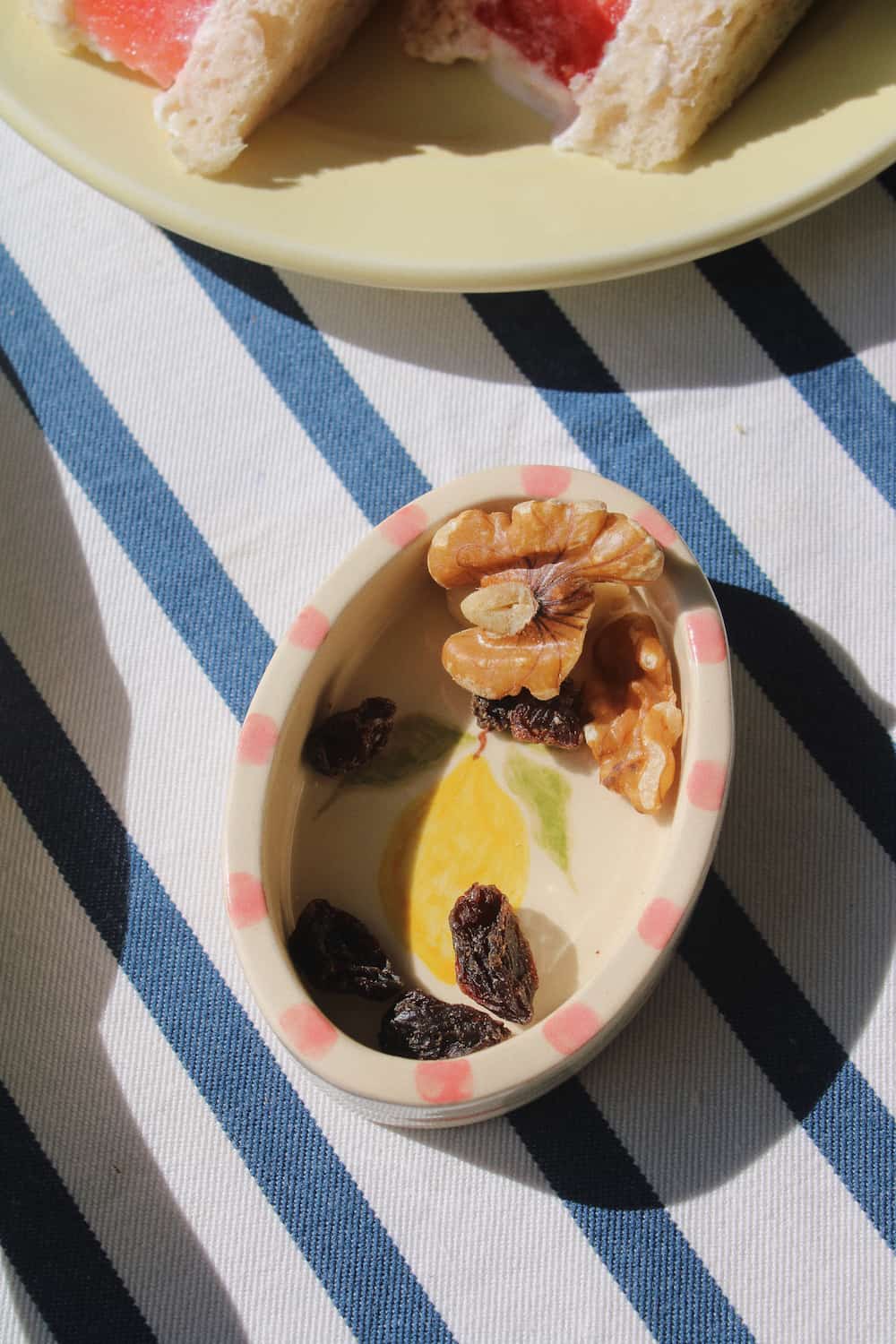 A small oval ceramic dish with pink polka dots around the rim, containing walnuts, raisins, and a yellow lemon slice, placed on a blue and white striped tablecloth with a plate of bread partially visible in the background.