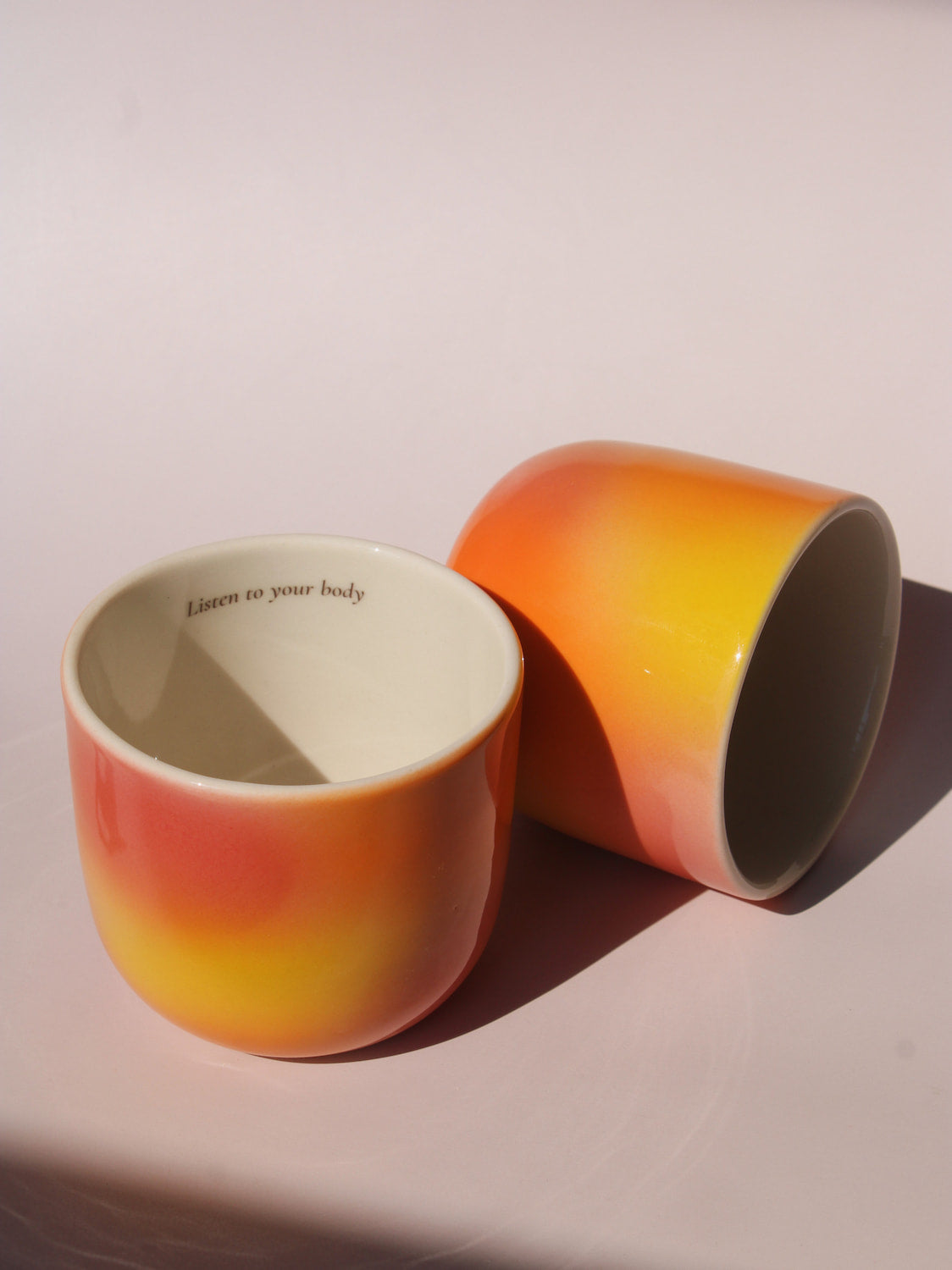 Gradient ceramic mugs in warm orange yellow and pink tones with "Listen to your body" printed inside the rim, handmade in small batches — Listen to Your Body limited edition ceramic mug bladder cancer awareness collection