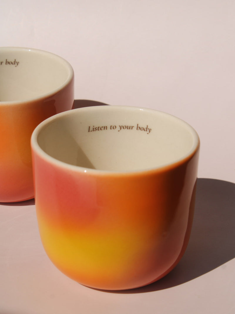 Gradient ceramic mugs in warm orange yellow and pink tones with "Listen to your body" printed inside the rim, handmade in small batches — Listen to Your Body limited edition ceramic mug bladder cancer awareness collection