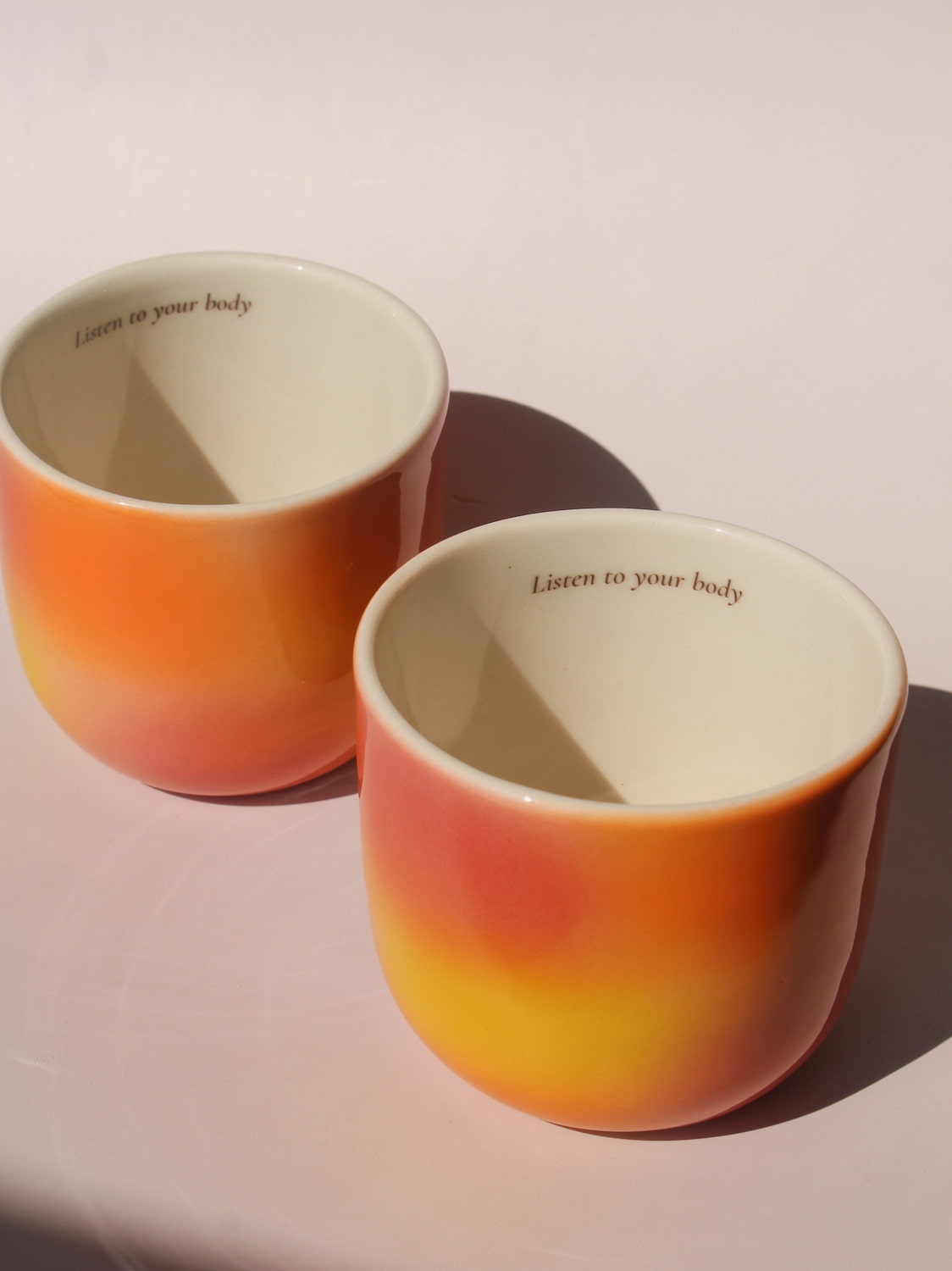 Gradient ceramic mugs in warm orange yellow and pink tones with "Listen to your body" printed inside the rim, handmade in small batches — Listen to Your Body limited edition ceramic mug bladder cancer awareness collection
