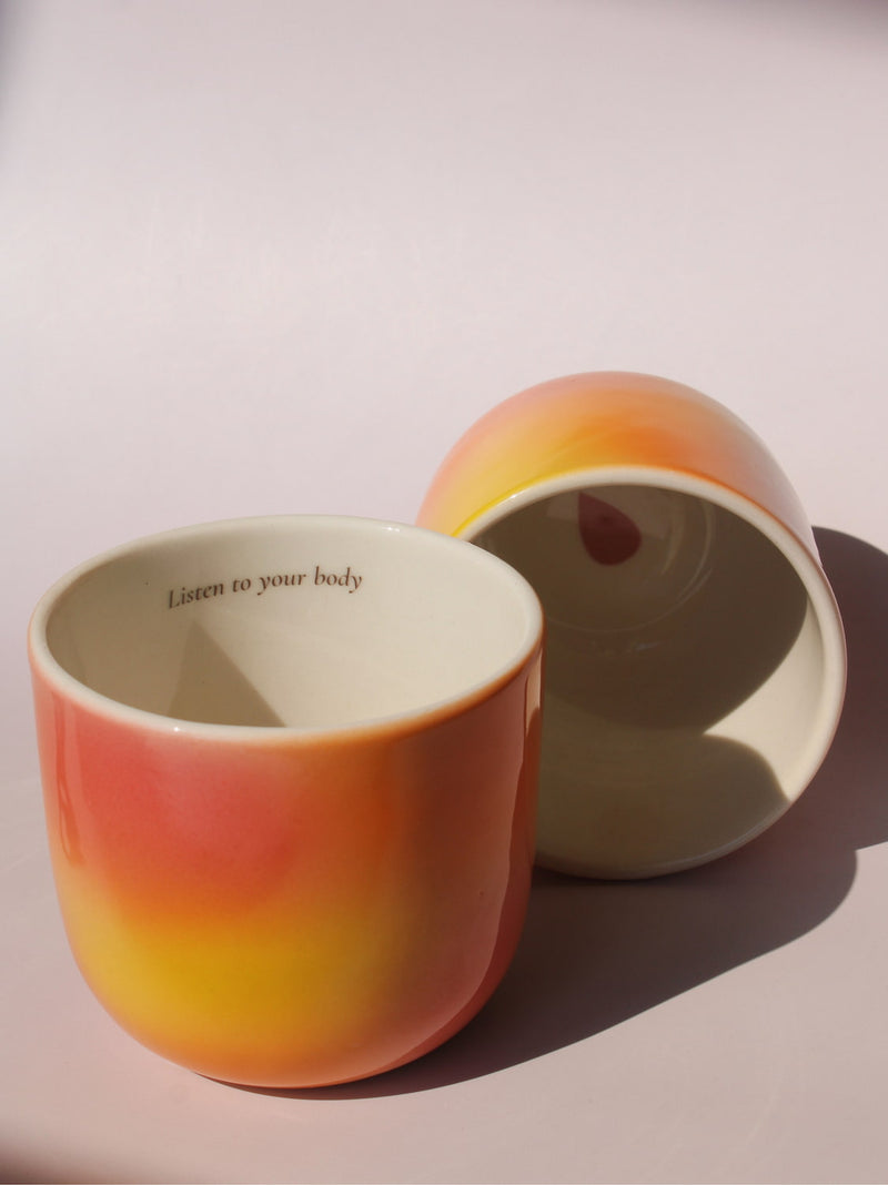 Gradient ceramic mugs in warm orange yellow and pink tones with "Listen to your body" printed inside the rim, handmade in small batches — Listen to Your Body limited edition ceramic mug bladder cancer awareness collection