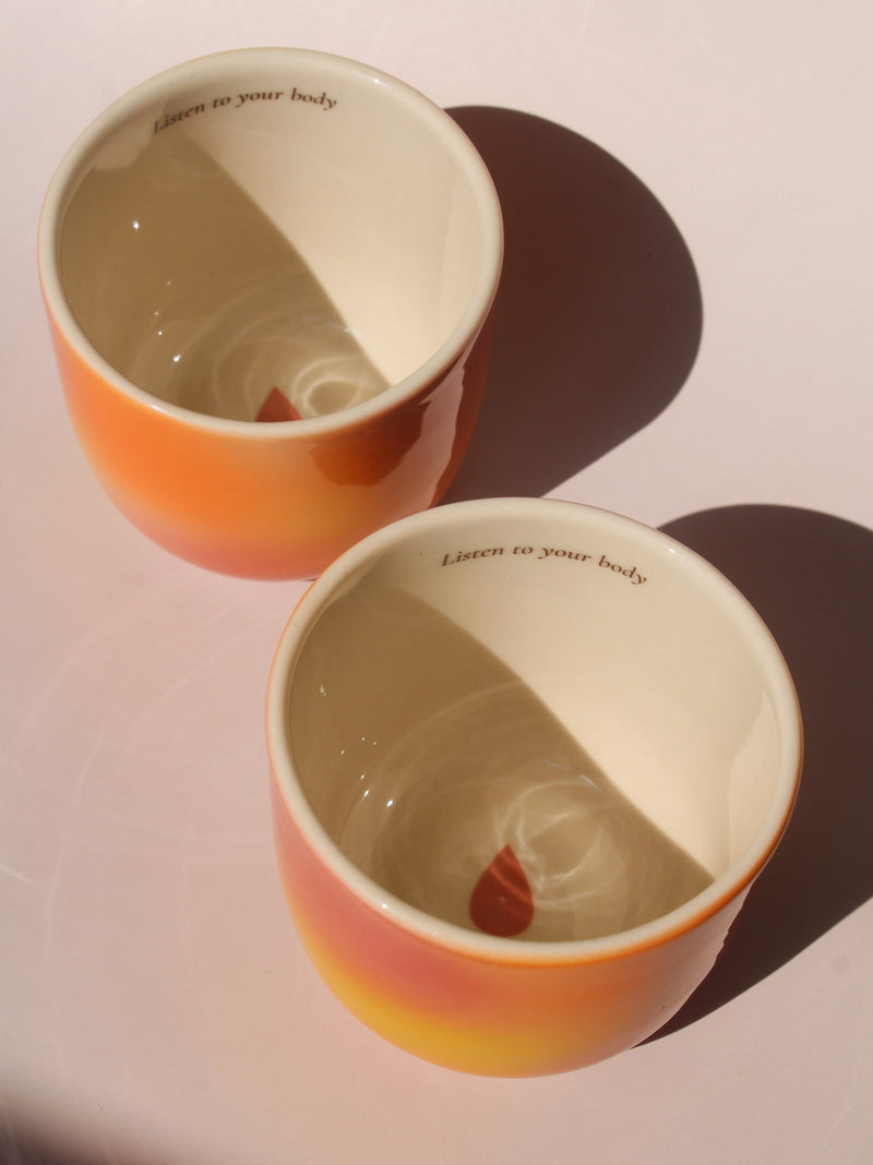 Gradient ceramic mugs in warm orange yellow and pink tones with "Listen to your body" printed inside the rim, handmade in small batches — Listen to Your Body limited edition ceramic mug bladder cancer awareness collection
