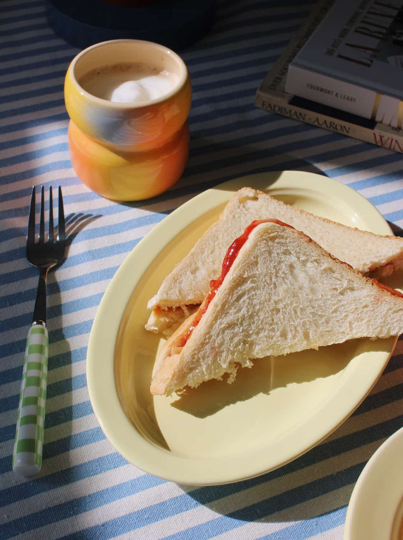 Triangular sandwich on a yellow plate with a striped tablecloth, fork, and cup in the background.