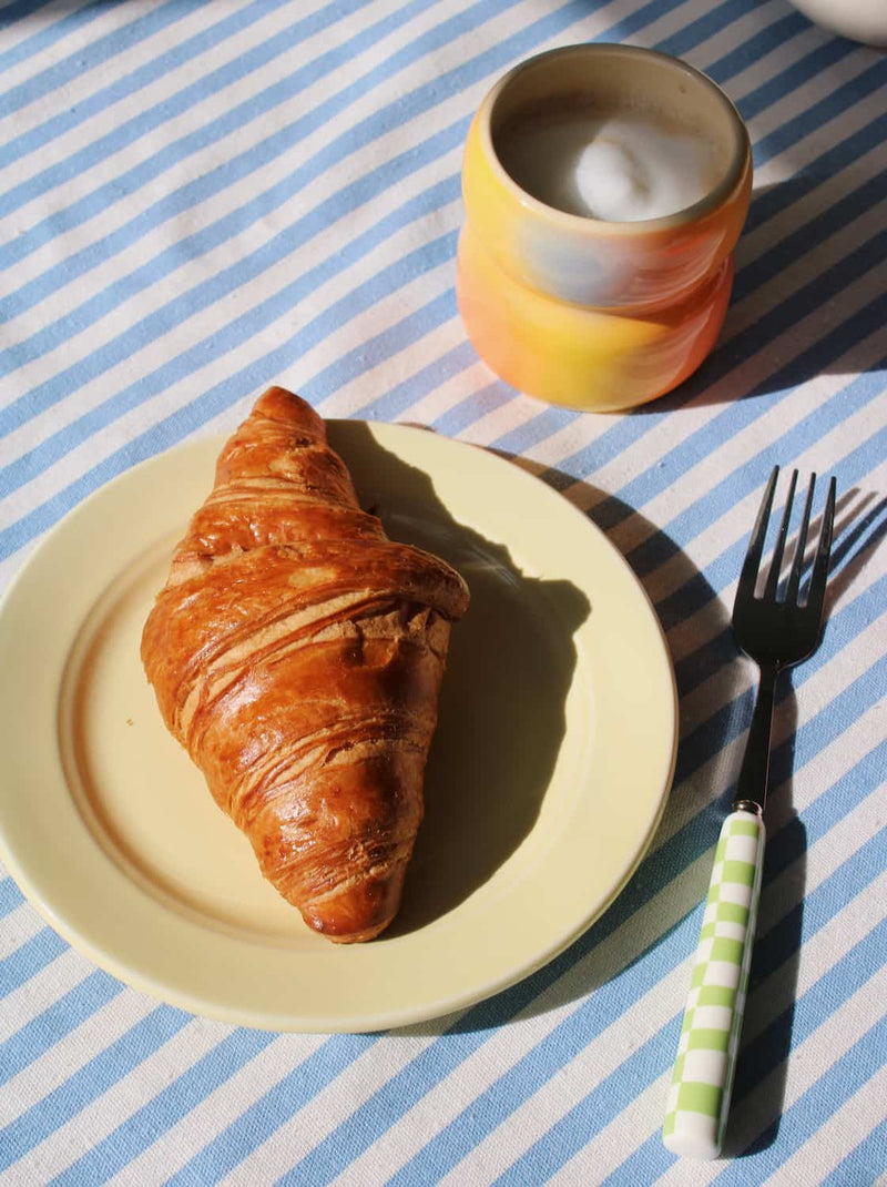 Croissant on yellow Lumi plate with a cup of coffee and a fork on a striped tablecloth