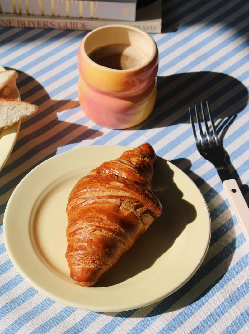 Croissant on yellow Lumi plate with a cup of coffee and a fork on a striped tablecloth