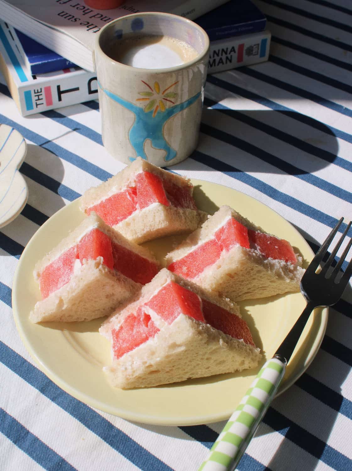 Four triangular sandwich slices filled with red watermelon and white cream on a pale yellow plate, set on a blue-and-white striped tablecloth with a fork, a coffee mug, and books in the background.
