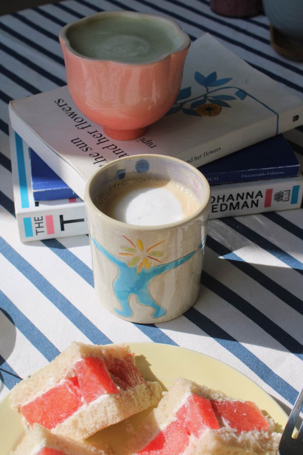 A cozy tabletop scene with a ceramic mug filled with a frothy drink in the foreground, a pink bowl behind it on top of books, and a plate of fruit slices in the lower corner, all set on a blue-and-white striped cloth.