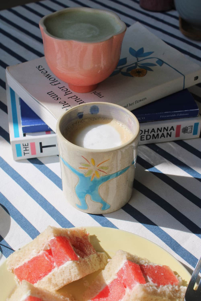 A cozy tabletop scene with a ceramic mug filled with a frothy drink in the foreground, a pink bowl behind it on top of books, and a plate of fruit slices in the lower corner, all set on a blue-and-white striped cloth.