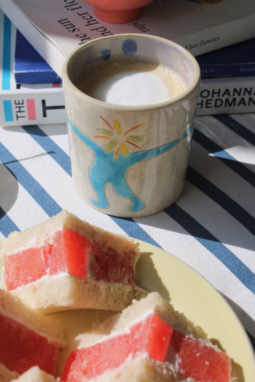 A beige ceramic mug with a blue and yellow sunburst design, filled with a light brown beverage, set on a blue and white striped tablecloth next to a plate of layered sponge cake with red fruit filling. Books are visible in the background.