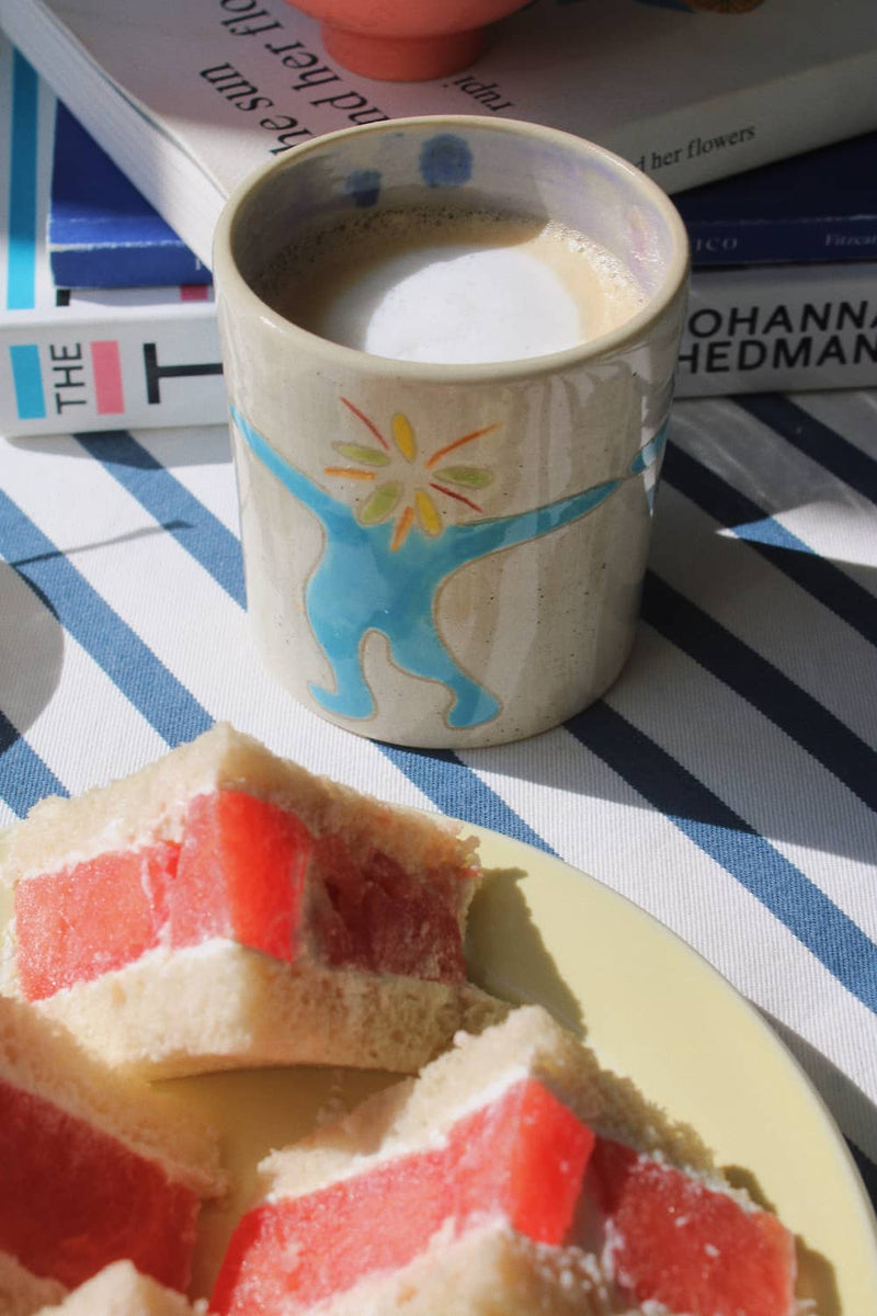 A beige ceramic mug with a blue and yellow sunburst design, filled with a light brown beverage, set on a blue and white striped tablecloth next to a plate of layered sponge cake with red fruit filling. Books are visible in the background.
