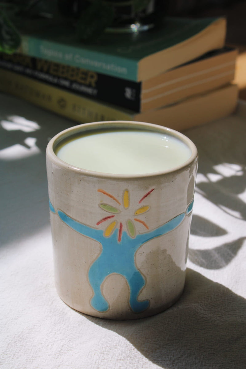 A handmade ceramic mug with a hand-painted blue figure and colorful sunburst design, filled with milk and set on a tablecloth with a stack of books in the background.