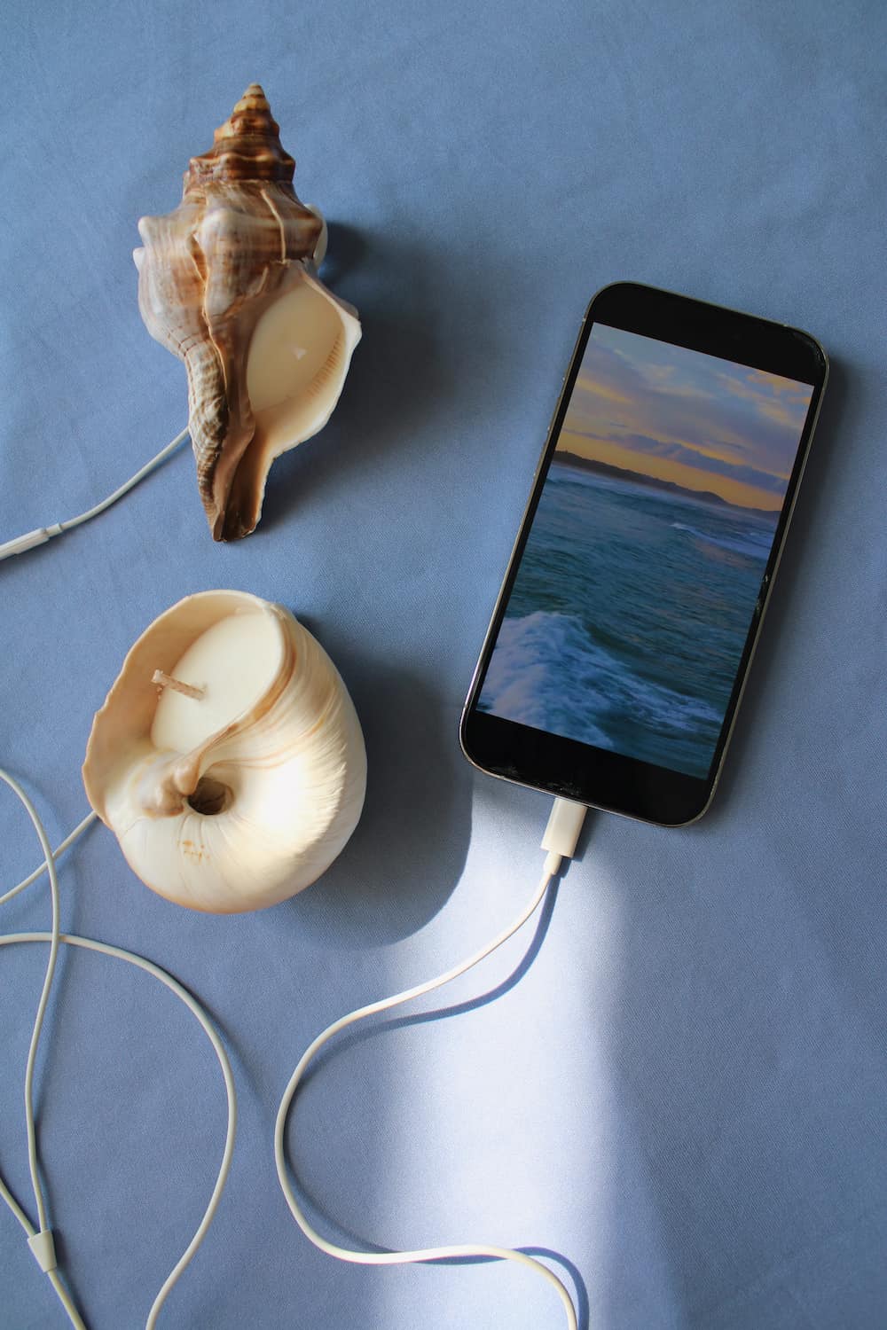 A smartphone lying on a blue surface, connected to white wired earphones, with two seashells placed nearby. The phone screen shows an ocean sunset scene.