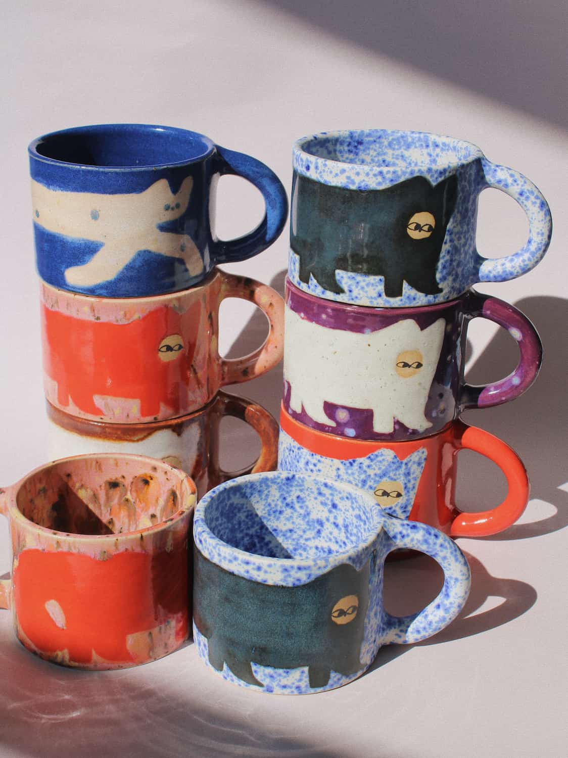 A set of handmade ceramic mugs stacked in two groups, featuring a hand-painted black cat design on speckled blue, orange, purple, and white glazes, with one mug showing a raw, unglazed interior.