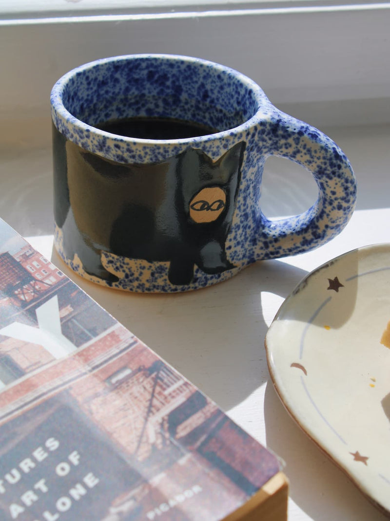 Midnight Cat ceramic mug with blue speckled rim and cat motif styled with books on windowsill