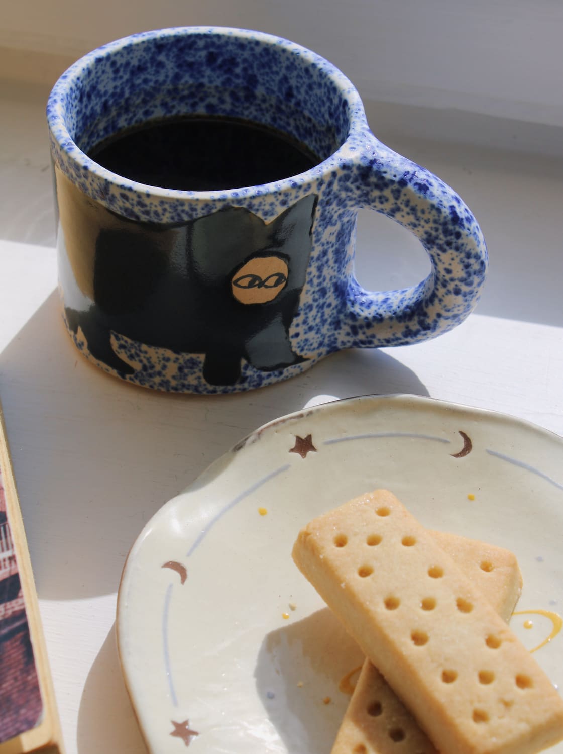 A speckled blue and white ceramic mug filled with black coffee, featuring a black cat illustration with a gold eye, set next to a plate of shortbread cookies on a white surface.