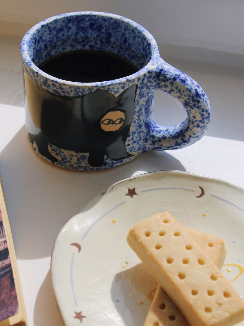 A speckled blue and white ceramic mug filled with black coffee, featuring a black cat illustration with a gold eye, set next to a plate of shortbread cookies on a white surface.