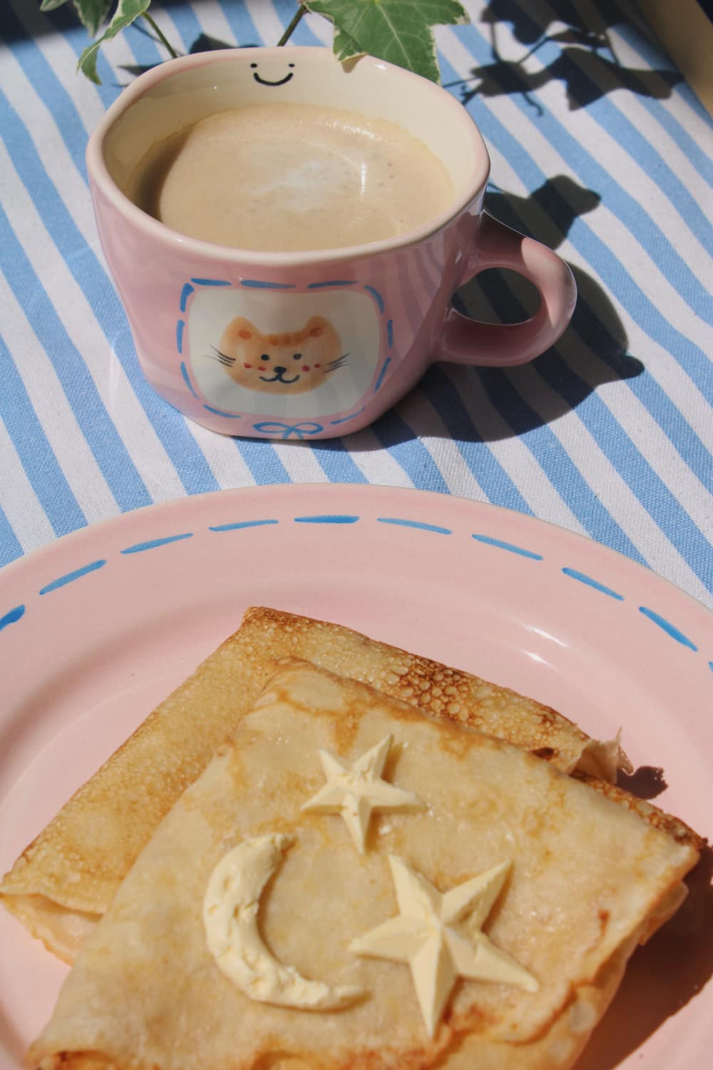 Mimi Ceramic Mug filled with coffee beside the Mimi Ceramic Plate styled with a buttered crepe on a blue striped surface, with ivy in the background.