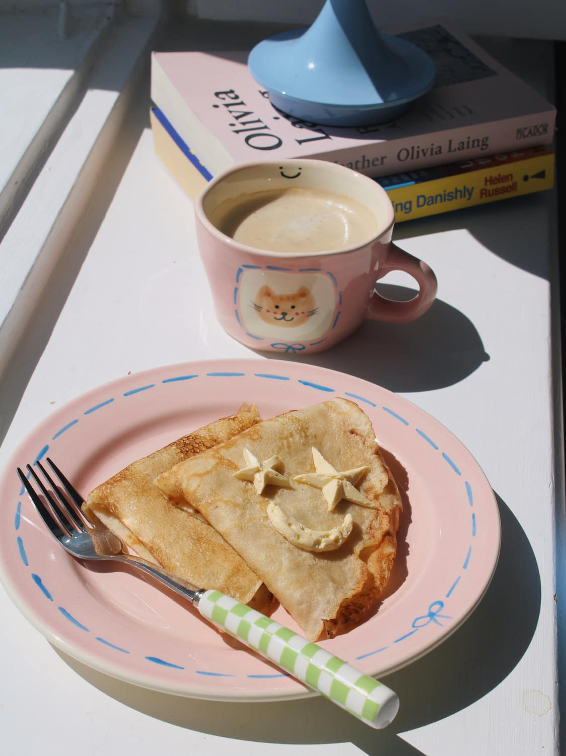 Mimi Ceramic Mug filled with coffee beside the Mimi Ceramic Plate styled with a buttered crepe on a blue striped surface, with ivy in the background.