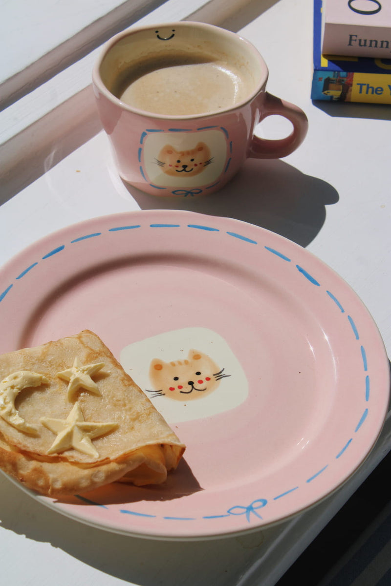 Mimi Ceramic Mug filled with coffee beside the Mimi Ceramic Plate styled with a buttered crepe on a blue striped surface, with ivy in the background.