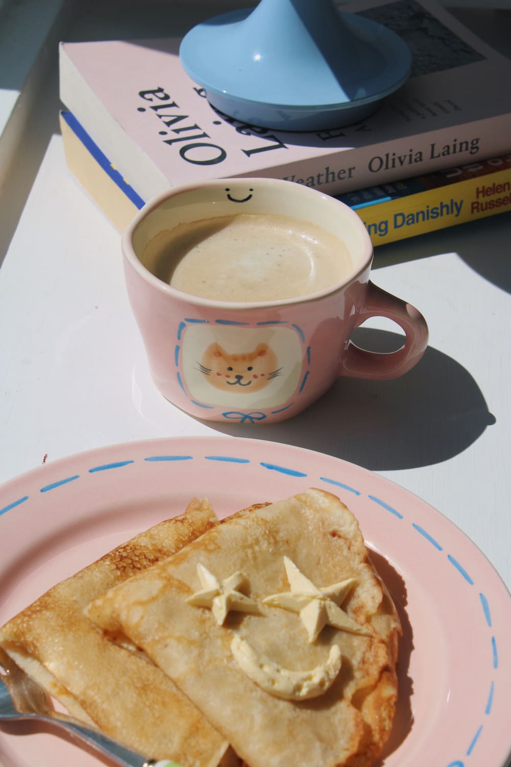 Mimi Ceramic Mug filled with coffee beside the Mimi Ceramic Plate styled with a buttered crepe on a blue striped surface, with ivy in the background.