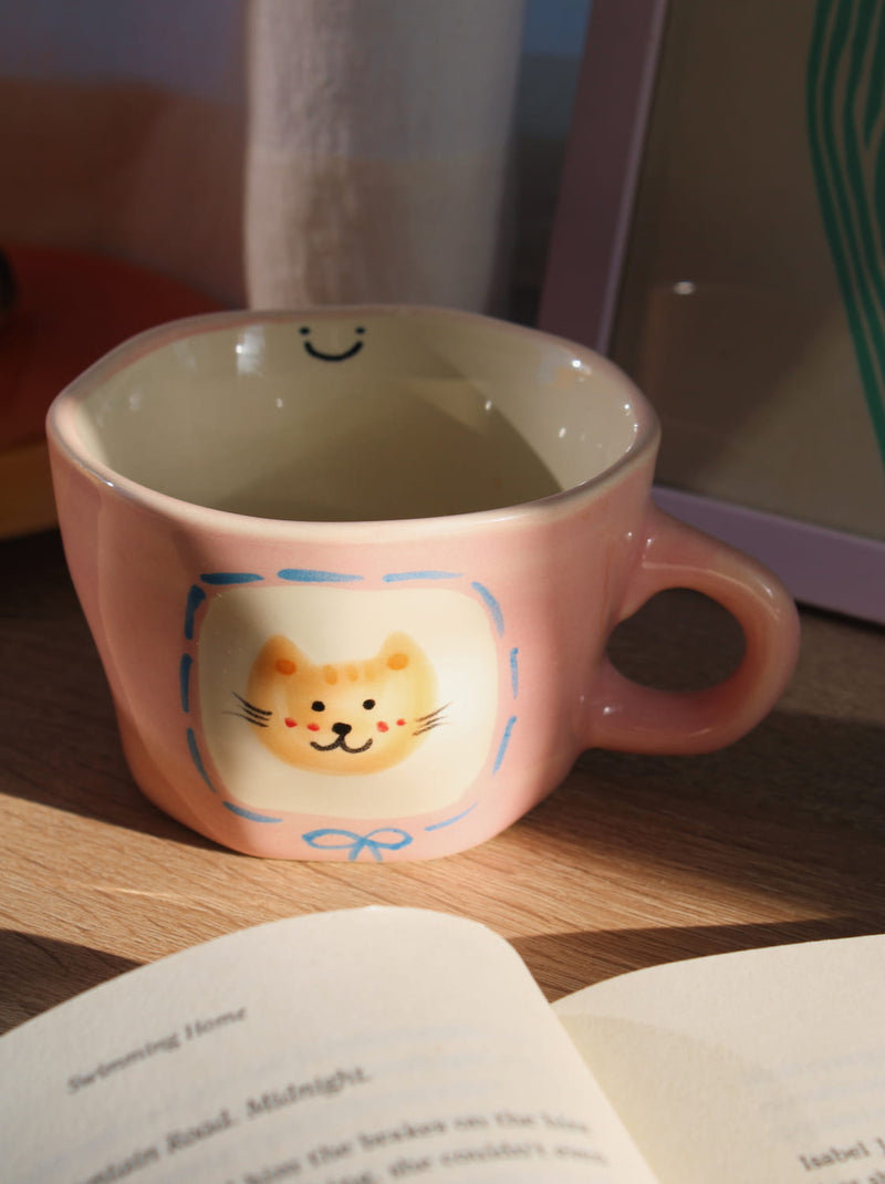 Pink mug with a cat design on a wooden surface next to an open book