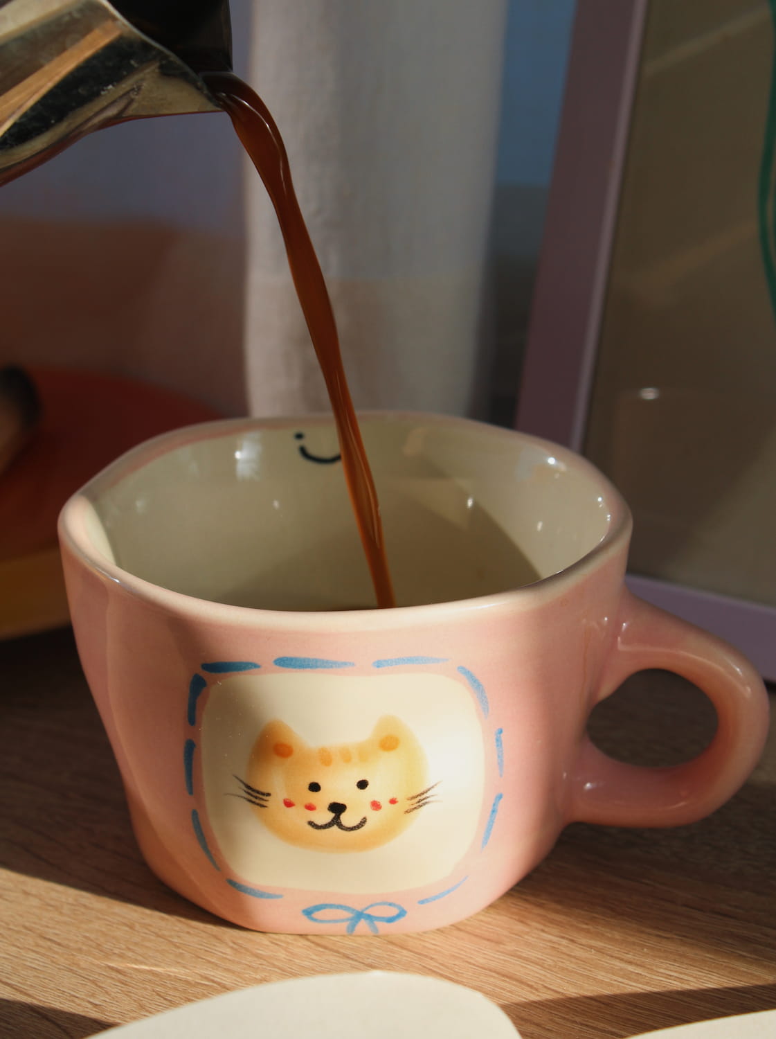 Pink mug with a cat design on a wooden surface next to an open book