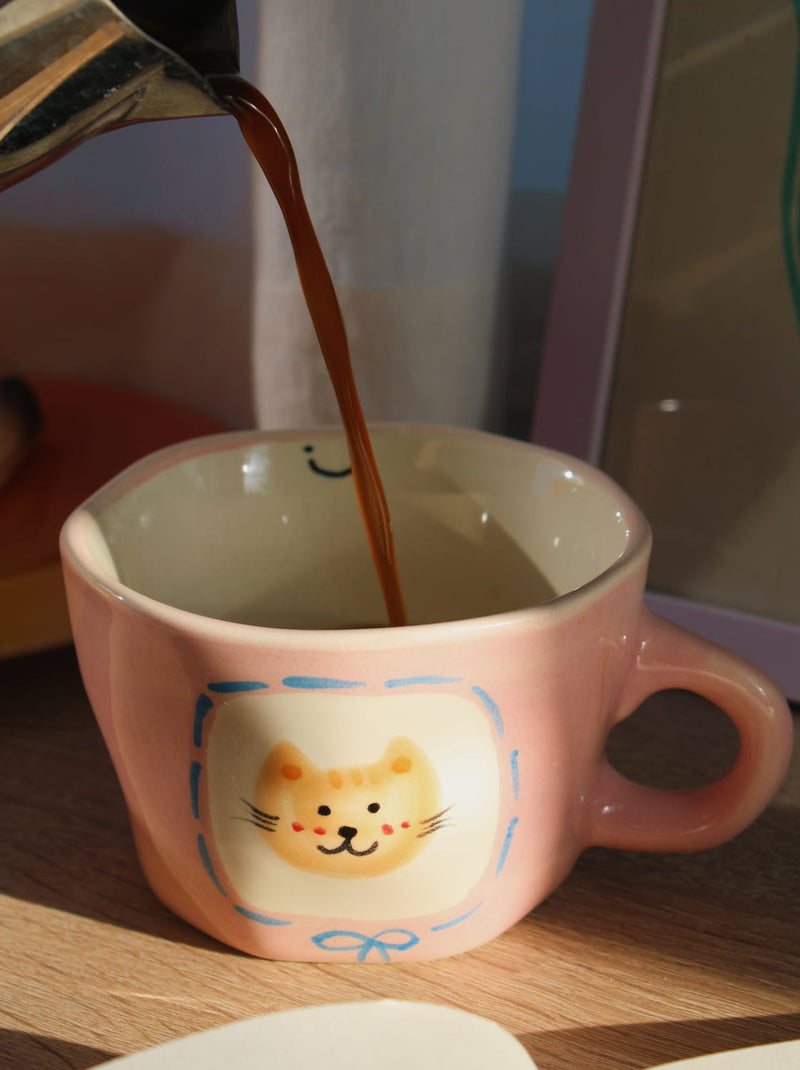 Pink mug with a cat design on a wooden surface next to an open book