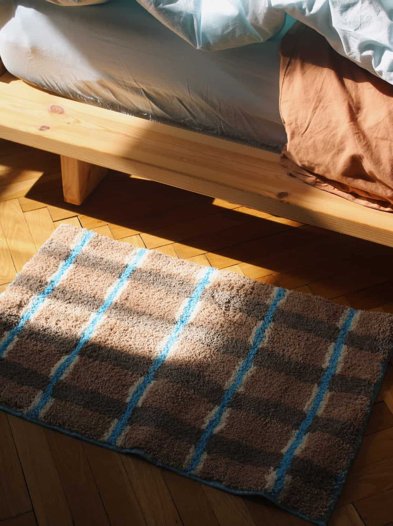 Checkered rug with blue lines on a wooden floor next to a bed.