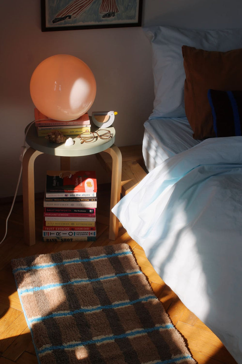 Bedroom scene with a nightstand, books, plaid rug on the floor.
