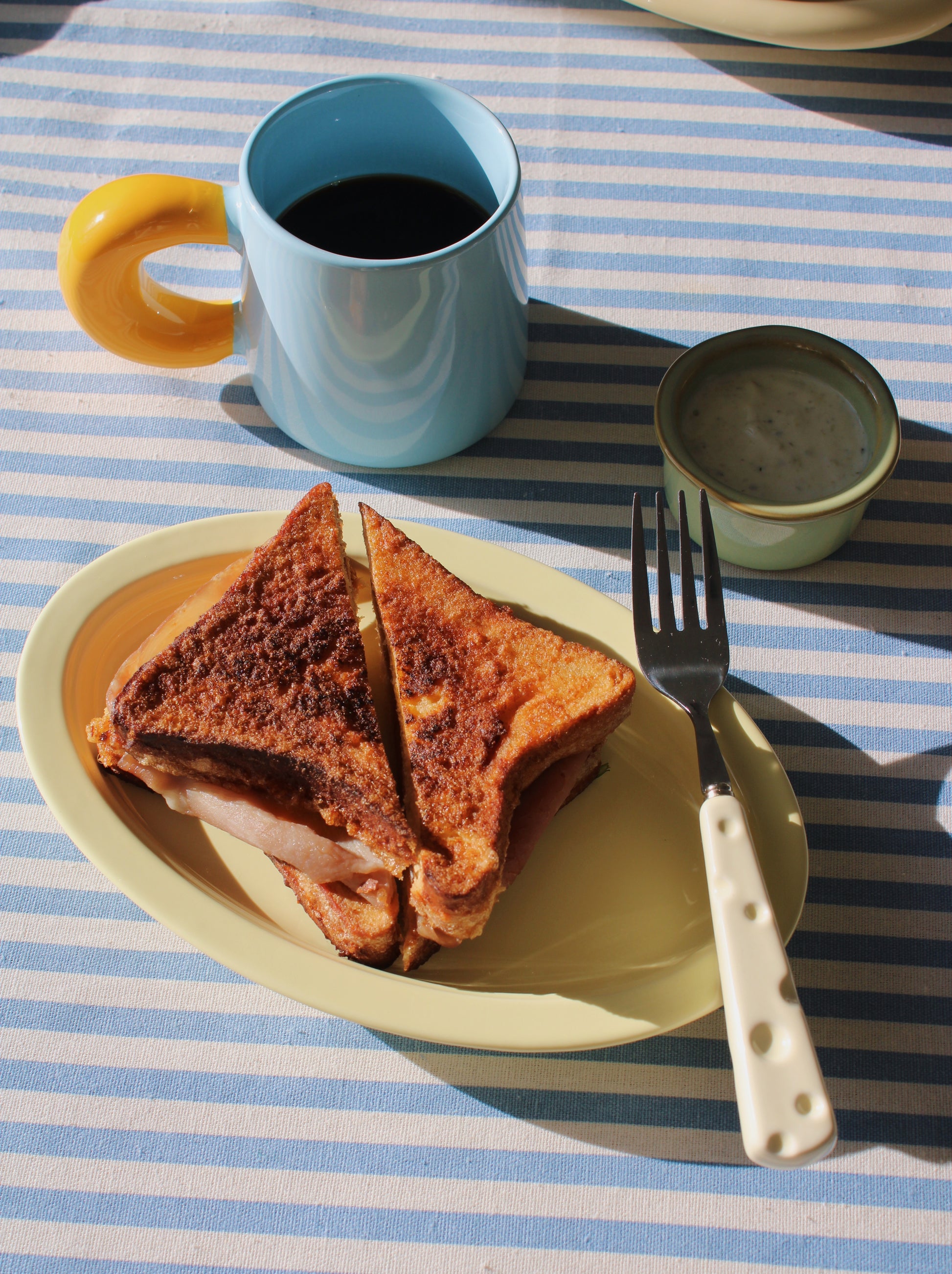 Toasted sandwich on a plate with a cup of coffee and a fork on a striped tablecloth.