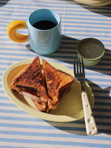 Toasted sandwich on a plate with a cup of coffee and a fork on a striped tablecloth.