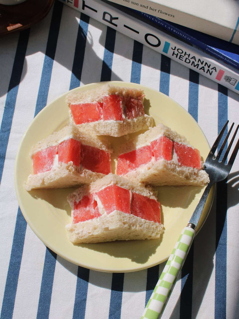 Four triangular sandwich slices filled with bright red fruit, arranged on a pale yellow plate with a fork on a blue-and-white striped tablecloth, with books in the background.