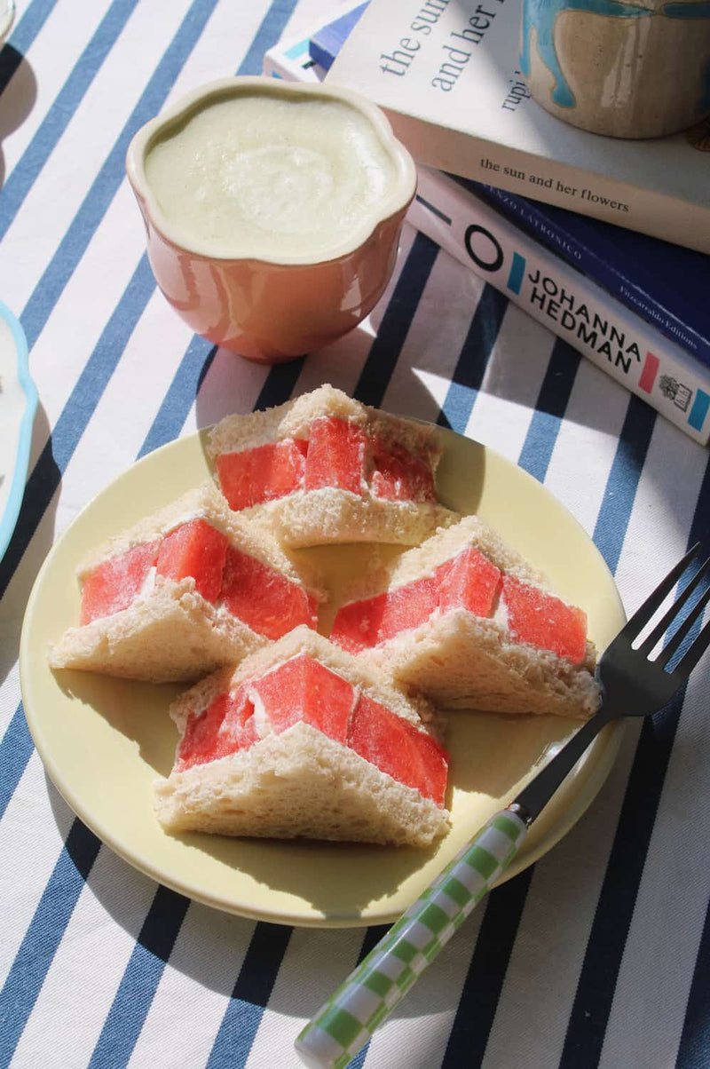 A styled breakfast scene with four triangular sandwich slices filled with red fruit, served on a pale yellow plate on a blue-and-white striped tablecloth. A pink cup of creamy drink, stacked books including one titled 'JOHANNA HEDMAN', and part of another mug are visible in the background.