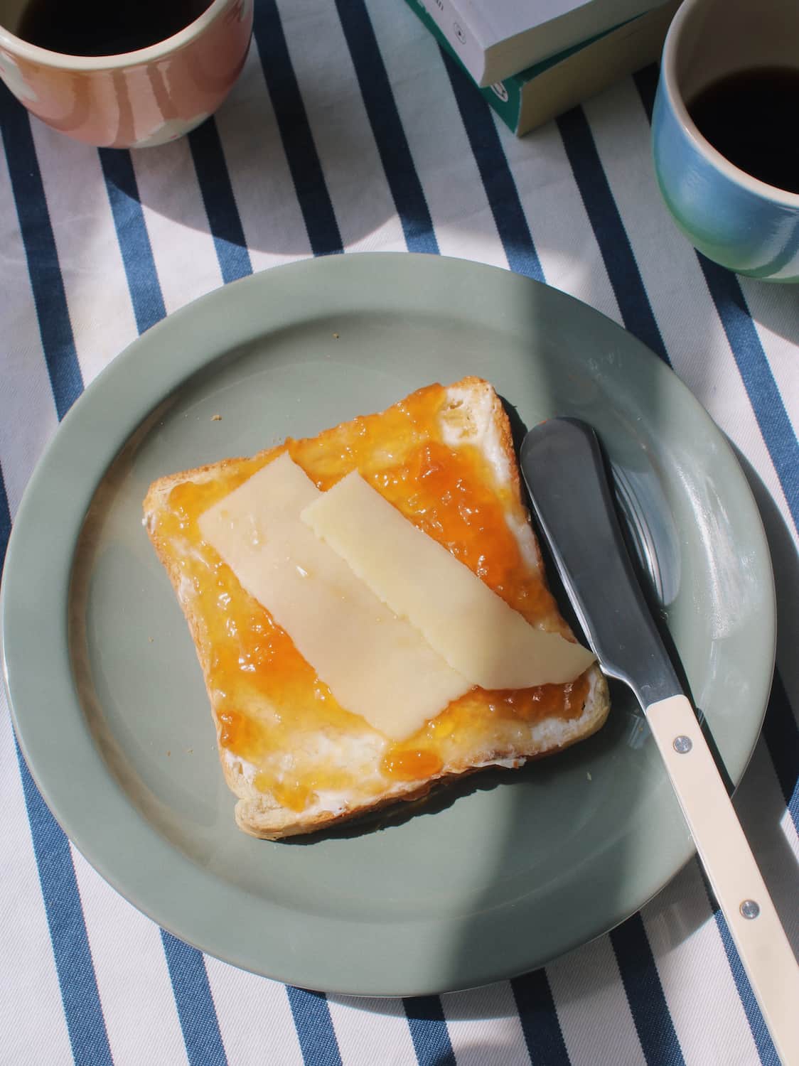 A slice of toast spread with orange jam and topped with two slices of white cheese on a light green plate, with a butter knife beside it on a blue-and-white striped tablecloth. Two mugs and a book are partially visible in the background.