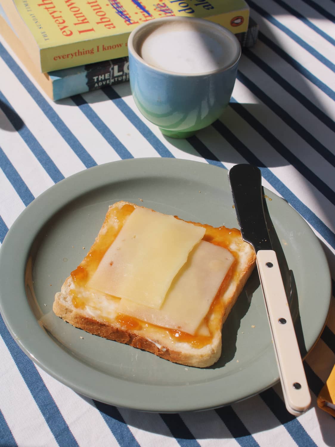 A slice of toast with cheese and orange spread on a light green plate, set on a blue-and-white striped tablecloth next to a knife, with a cup and books in the background.
