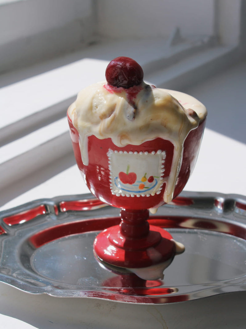Red ceramic goblet with ice cream sundae with a cherry on top on a metal stand.