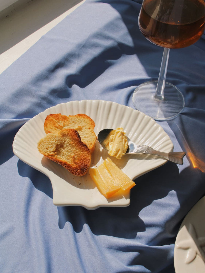 Pisca Spoon with fish tail detail resting on the Calista Ceramic Shell Plate, styled with toasted bread, butter and cheese alongside a glass of rosé wine on a blue fabric surface in natural sunlight.