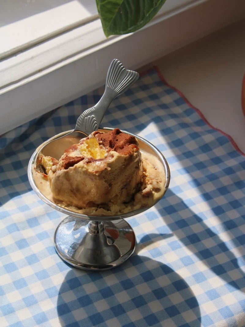 Pisca Spoon with fish tail detail resting in a silver pedestal ice cream dish filled with tiramisu, on a blue gingham surface with a terracotta plant pot by a sunny window.