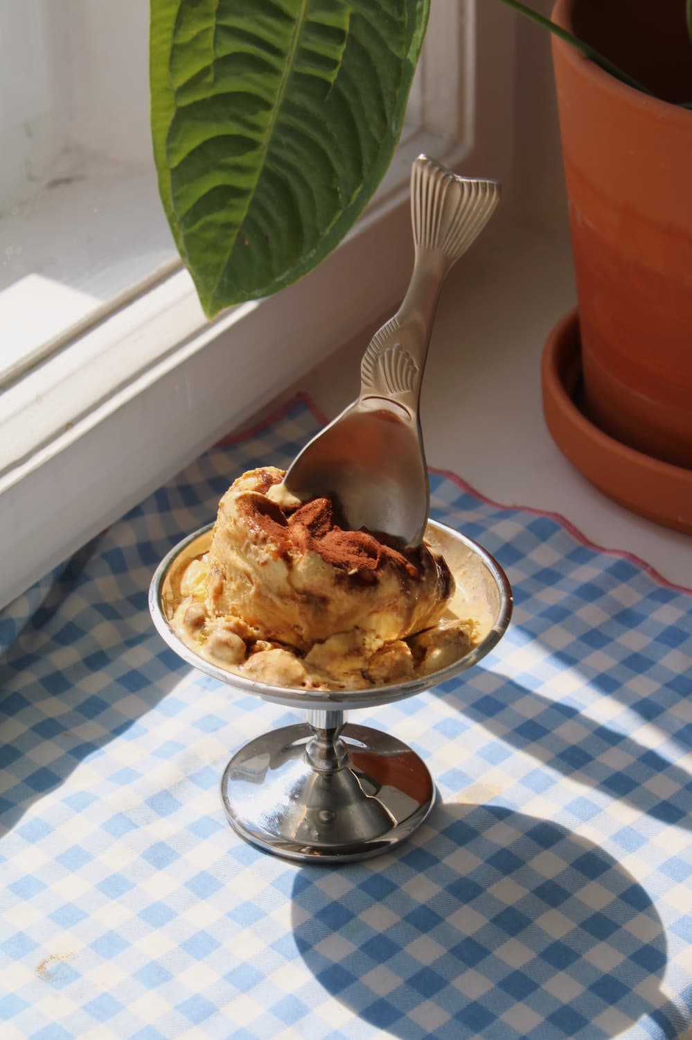 Pisca Spoon with fish tail detail resting in a silver pedestal ice cream dish filled with tiramisu, on a blue gingham surface with a terracotta plant pot by a sunny window.