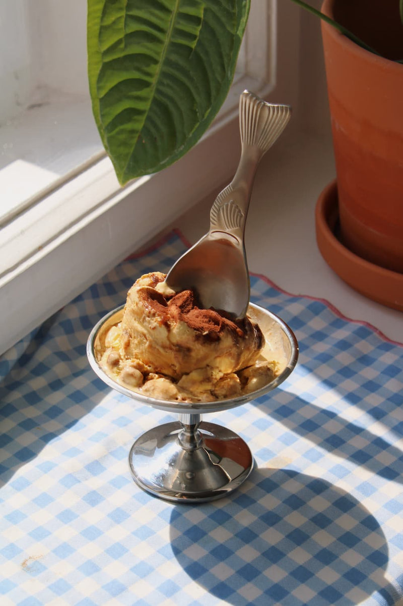 Pisca Spoon with fish tail detail resting in a silver pedestal ice cream dish filled with tiramisu, on a blue gingham surface with a terracotta plant pot by a sunny window.