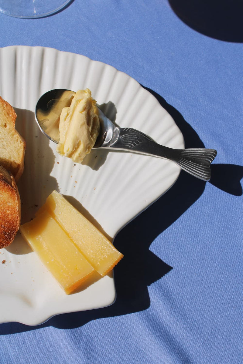 Pisca Spoon with fish tail detail resting on the Calista Ceramic Shell Plate, styled with toasted bread, butter and cheese alongside a glass of rosé wine on a blue fabric surface in natural sunlight.
