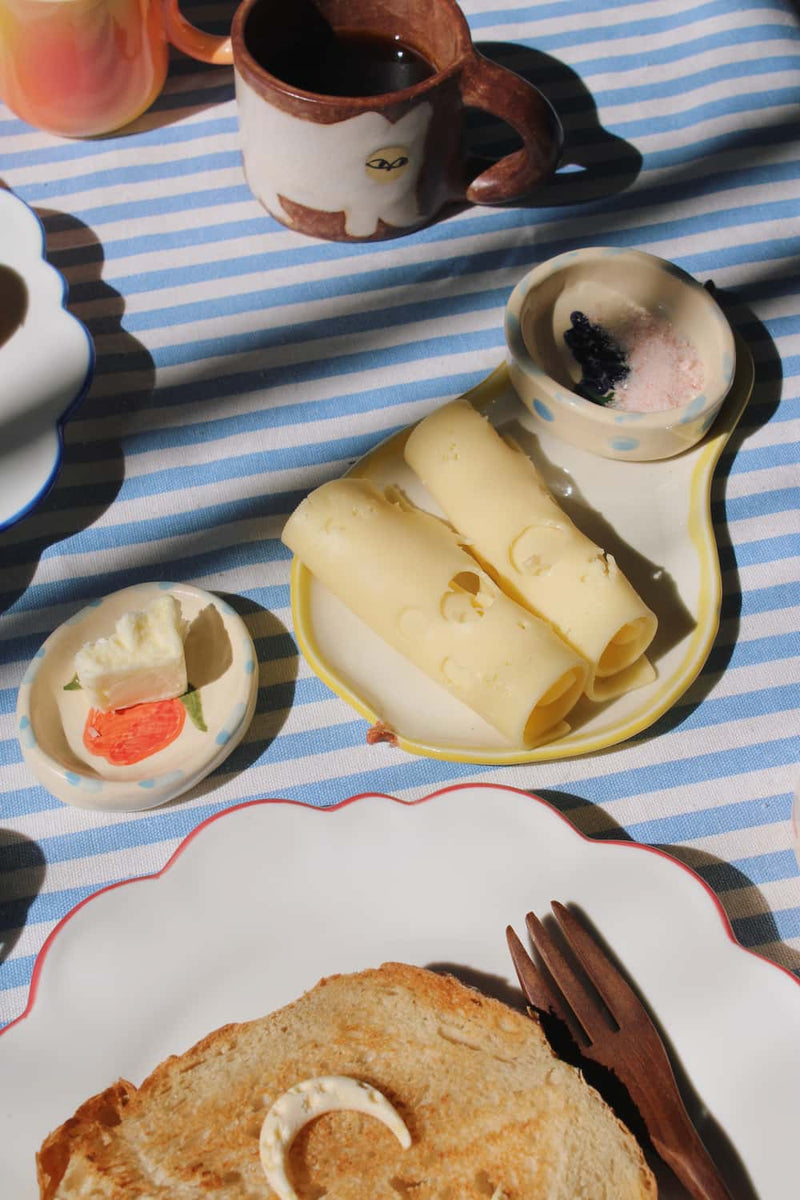 Breakfast setting with toast, rolled cheese on handmade ceramic trinket, and coffee on a striped tablecloth.