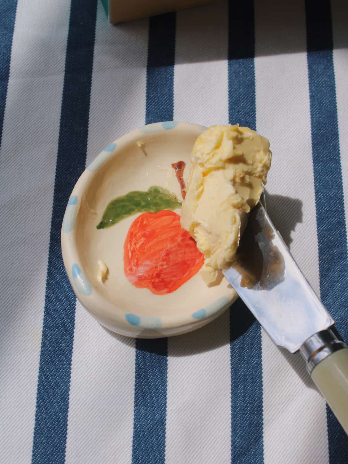 A small hand-painted ceramic dish with a painted red apple and green leaf, holding a knife with pale yellow butter on a blue and white striped tablecloth.
