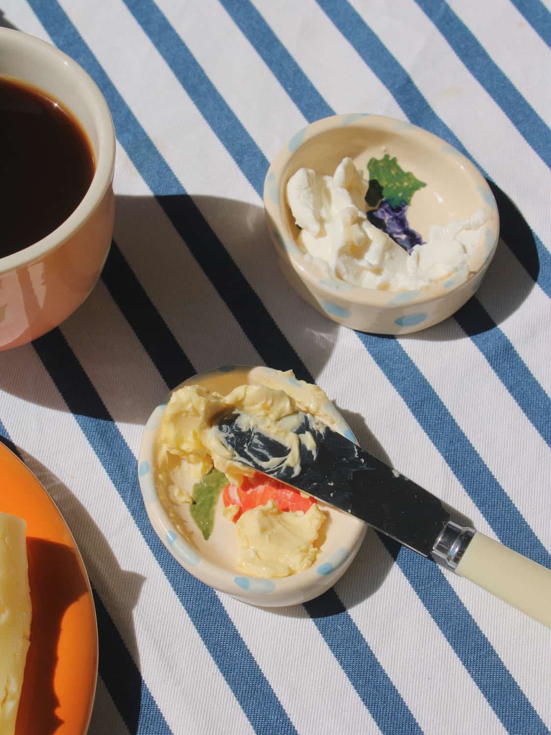 Two small cream-colored bowls with blue polka dots sit on a blue and white striped tablecloth. One bowl contains whipped cream with a green leaf garnish, and the other contains butter spread with a knife, set next to a cup of coffee.