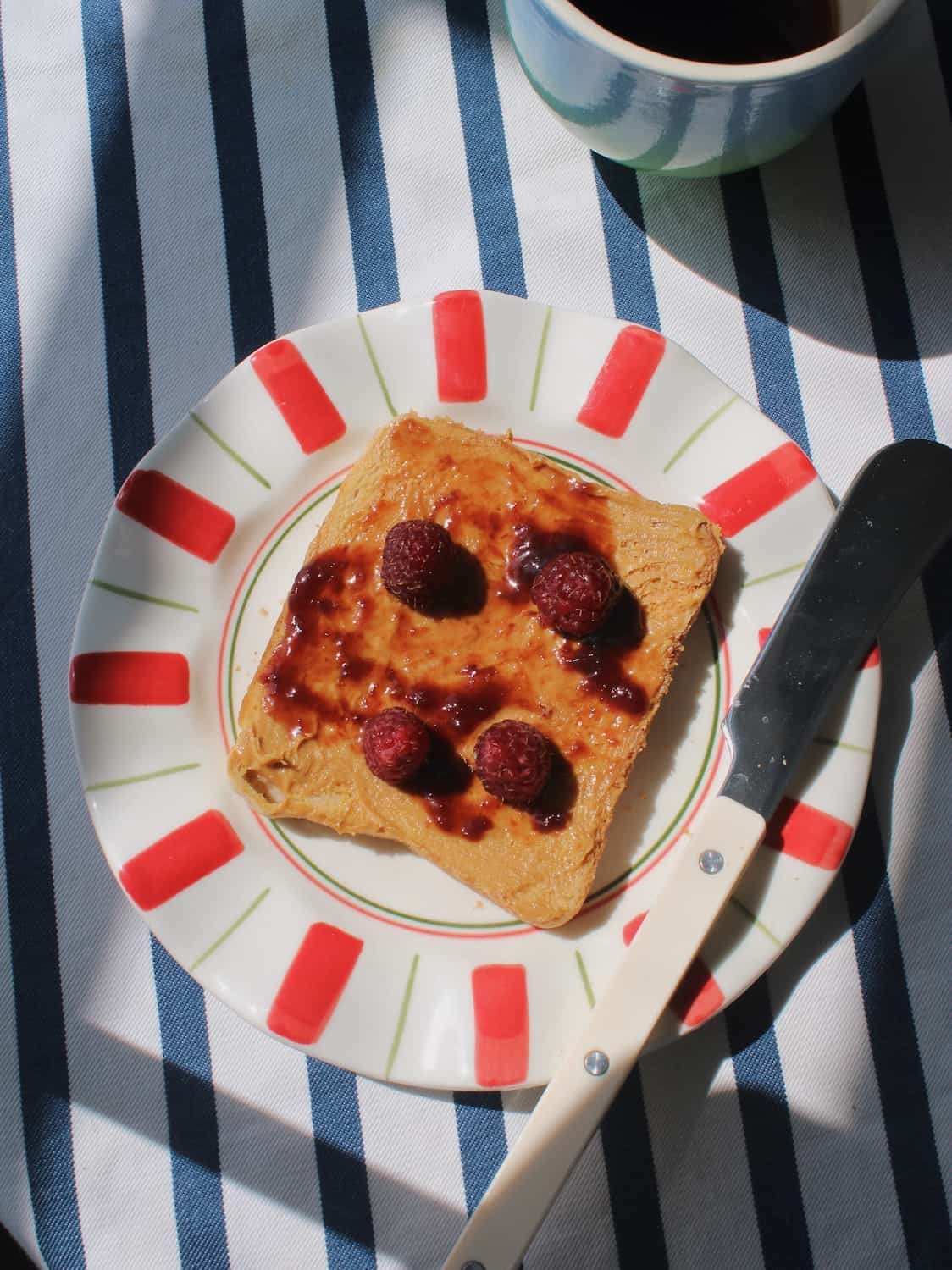 A slice of toast topped with jam and three raspberries on a white plate with red and green stripes, set on a blue and white striped tablecloth with a knife and a cup nearby.