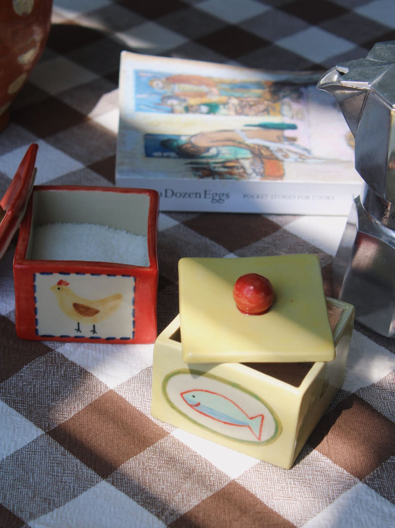 Sardina Ceramic Box in pale yellow and Henley Trinket Box in red with hand-painted chicken illustration, styled open together on a brown gingham surface with a Bialetti moka pot and stacked books in the background.