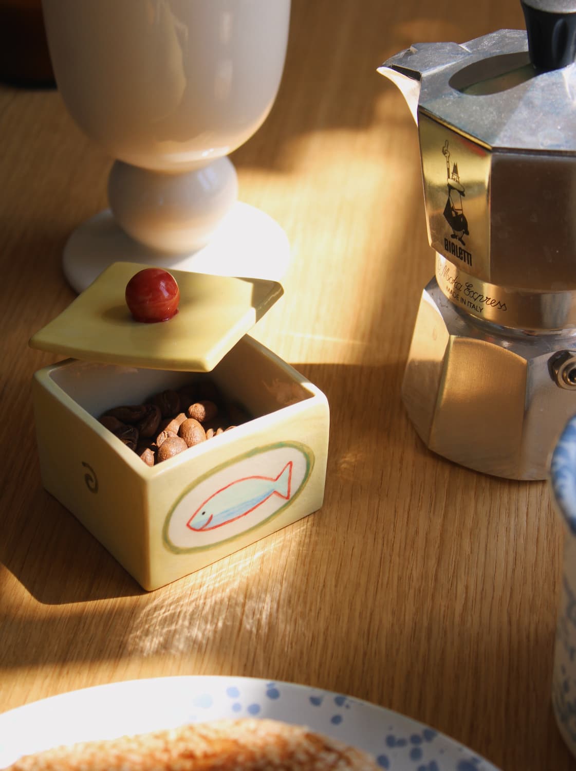 Sardina Ceramic Box in pale yellow with hand-painted sardine illustration, open and filled with coffee beans beside a Bialetti moka pot and white egg cup on a wooden surface in warm natural light.